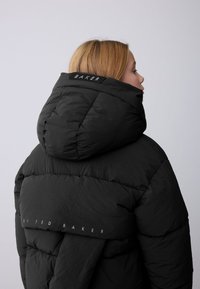 Zwarte donzen jas met capuchon, met een gewatteerde textuur. "BAKER" logo in het wit op de capuchon, en "by TED BAKER" op de achterkant.