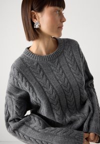 Pull en maille gris avec un motif de torsades, col rond et épaules tombantes pour une coupe décontractée. Texture douce et poids moyen.