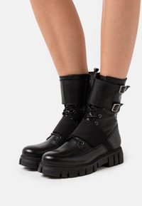 Bottes courtes en cuir noir à finition mate, avec un bord rembourré, des boucles ajustables et des semelles épaisses en caoutchouc. Modèle à lacets.