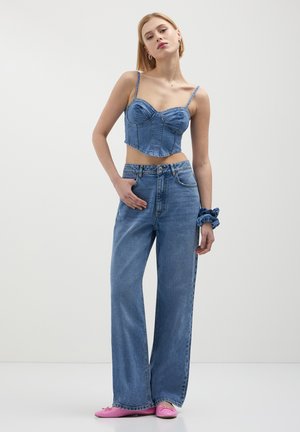 Top corsetto in denim a maniche sottili, accorciato, abbinato a jeans in denim a gamba larga. Colore blu chiaro, cuciture visibili e accessori includono un elastico per capelli.