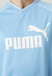Lyseblå sportstrøje lavet af tekstureret stof. Har et hvidt PUMA-logo og tydelig skrift. V-halsdesign med kontrastkant.