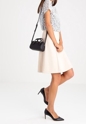 Femme portant un chemisier en dentelle blanche, une jupe beige, des escarpins noirs et portant un petit sac bandoulière noir avec des poignées.