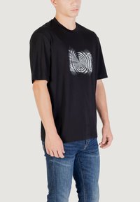 T-shirt in cotone nero con maniche corte, caratterizzato da una grafica circolare bianca sulla parte anteriore. Abbinato a jeans blu, con un taglio casual.