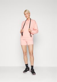 Rosa hettegenser med snor og frontlomme, kombinert med matchende shorts. Svarte treningssko og sokker fullfører antrekket.