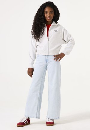 Lichtgrijze zip-up hoodie met een cropped ontwerp, logo op de linkerkant van de borst, gecombineerd met hoge taille lichtblauwe wijde jeans en rode schoenen.