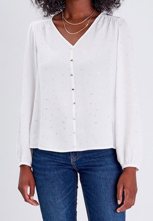 Femme portant un chemisier blanc à manches longues avec des motifs argentés subtils et un jean bleu, accessoirisée avec deux colliers en or.