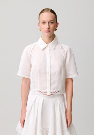 Mujer con cabello rubio en una coleta baja que lleva una camisa blanca de manga corta con detalles de encaje y una falda blanca a juego sobre un fondo gris.