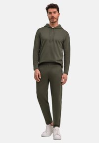 Olivgrüner Hoodie mit Kordelzugkapuze, kombiniert mit passenden, figurbetonten Hosen. Weiße Sneaker und ein strukturiertes Stoffdesign.