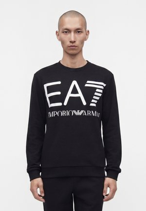 Homme aux cheveux courts portant un sweat-shirt noir avec un grand logo blanc "EA7 Emporio Armani" se tenant devant un fond blanc uni.