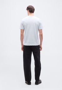 Άντρας που στέκεται με το πρόσωπο προς τα πίσω, φορώντας ανοιχτό γκρι μπλουζάκι, μαύρο τζιν και σκούρα παπούτσια, με φόντο απλό λευκό.