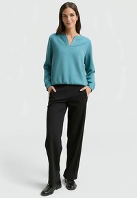 Maglione turchese con scollo a V e dettagli sottili, abbinato a pantaloni neri ampi e scarpe nere. Tessuto dalla texture liscia.