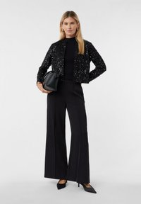 Kobieta w czarnej cekinowej kurtce, czarnych szerokich spodniach, czarnych szpiczastych szpilkach, trzymająca czarną torebkę na tle białym.