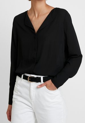 Person wearing a long-sleeve black blouse tucked into high-waisted white pants with a black belt, hand in pocket.