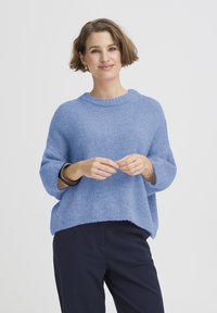 Pull en maille bleu clair avec une coupe d écontractée, manches larges et col côtelé. Porté avec un pantalon foncé, mettant en valeur un tissu doux et texturé.