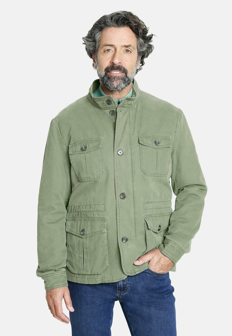 Hombre con cabello y barba rizados de sal y pimienta, vistiendo una chaqueta verde abotonada con bolsillos con solapa, jeans azules, de pie con una mano en el bolsillo.