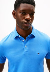 Blå pikéskjorte laget av teksturert stoff, med en plattform med tre knapper og en liten brodert logo på brystet. Kortermet.