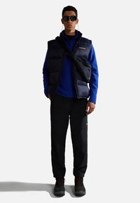 Marine puffer vest over een blauwe lange mouwen shirt, zwarte broek en donkere laarzen. Draagt een zonnebril en een schoudertas; de broek heeft zijzakken.