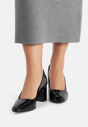 Czarne lakierowane szpilki, z zaokrąglonym noskiem, słupkowym obcasem i gładką powierzchnią. Połączone z szarą, sięgającą do kolan odzieżą.