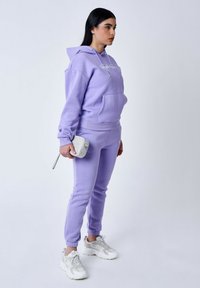 Paarse hoodie met voorzak en trekkoord, gecombineerd met taps toelopende joggers. Zilveren clutch, witte sneakers met textuurelementen.