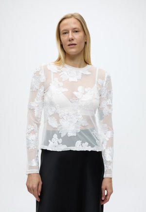 Woman wearing a sheer white floral embroidered long-sleeve top over a white bra, paired with a black skirt, standing against plain background.