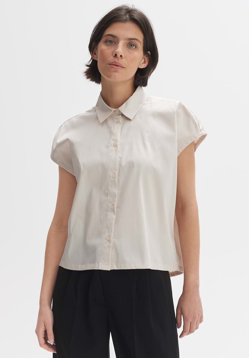 Short-sleeve, collarless blouse in light beige fabric with a smooth texture, featuring a button-down front and a relaxed fit.