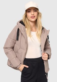 Polstret jakke i lys taupe med hætte, lynlås og sorte accenter. Har elastiske manchetter og sidelommer. Kombineret med en cremefarvet top.