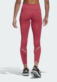 Taillierte, hochtaillierte Leggings in gedämpftem Rot mit Netzpaneelen und reflektierenden Akzenten an den Unterschenkeln. Kombiniert mit schwarzen Sportschuhen.