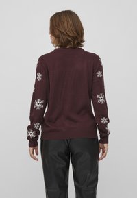 Burgundy stickad tröja med vita snöflinge-mönster på ärmarna, ribbade mudd och fåll, mjuk textur, avslappnad passform.