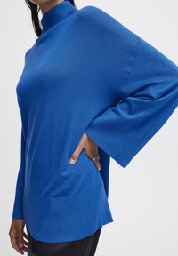 Pulover oversized de culoare albastră, cu o guleră înaltă și manșete largi. Țesătura este moale și netedă, având o textură ribbed la tiv.