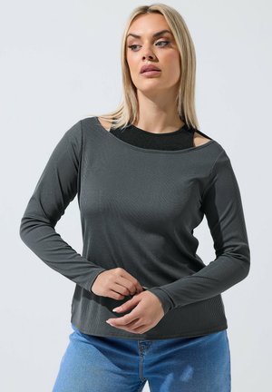 Kvinde med blond hår iført en mørkegrå langærmet top med udskårne skuldre og blå jeans, justerer ærmerne, mens hun kigger til siden.