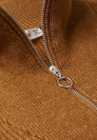 Brauner Strickpullover mit silbernem Reißverschluss, versehen mit einem strukturierten Stoff und einem Etikett in der Nähe des Kragens. Der Reißverschluss-Schieber hat einen runden Ring.