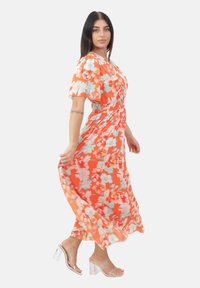 Robe maxi florale orange avec des fleurs bleu clair et blanches, manches courtes bouffantes, taille froncée, jupe fluide, associée à des talons transparents.