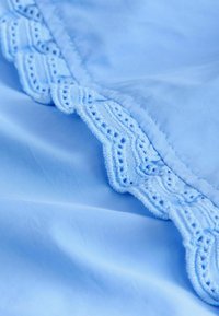 Tissu bleu clair avec une bordure en dentelle festonnée détaillée, présentant des motifs géométriques brodés le long du bord.