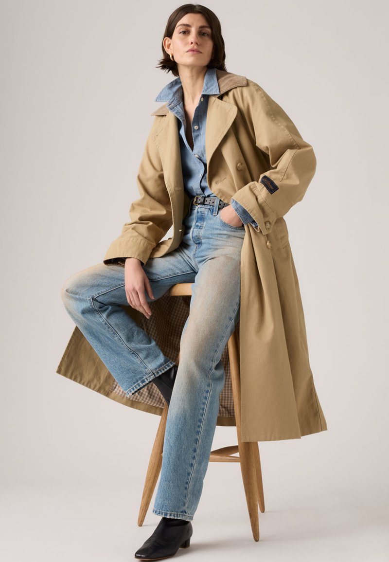Woman sitting on wooden stool wearing beige trench coat, blue denim shirt, light blue jeans, black ankle boots, and a black belt.