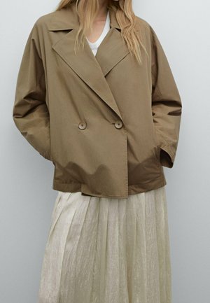 Woman wearing a loose, double-breasted, light brown jacket over a pleated off-white skirt, standing against a plain light background.