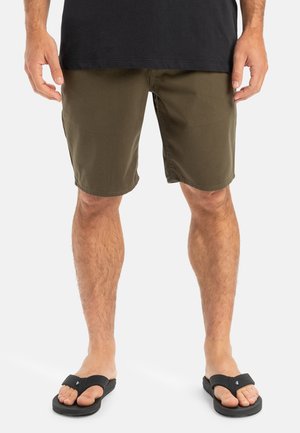 Khaki šortky vyrobené z hladkého materiálu, so strihom v rovnom strihu a vo voľnom strihu. V kombinácii s čiernymi šľapkami a tmavým tričkom.