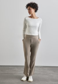 Maglietta bianca a maniche lunghe a costine, pantaloni beige sartoriali con tasche laterali e sneaker bianche. Il modello è in piedi contro uno sfondo grigio chiaro.