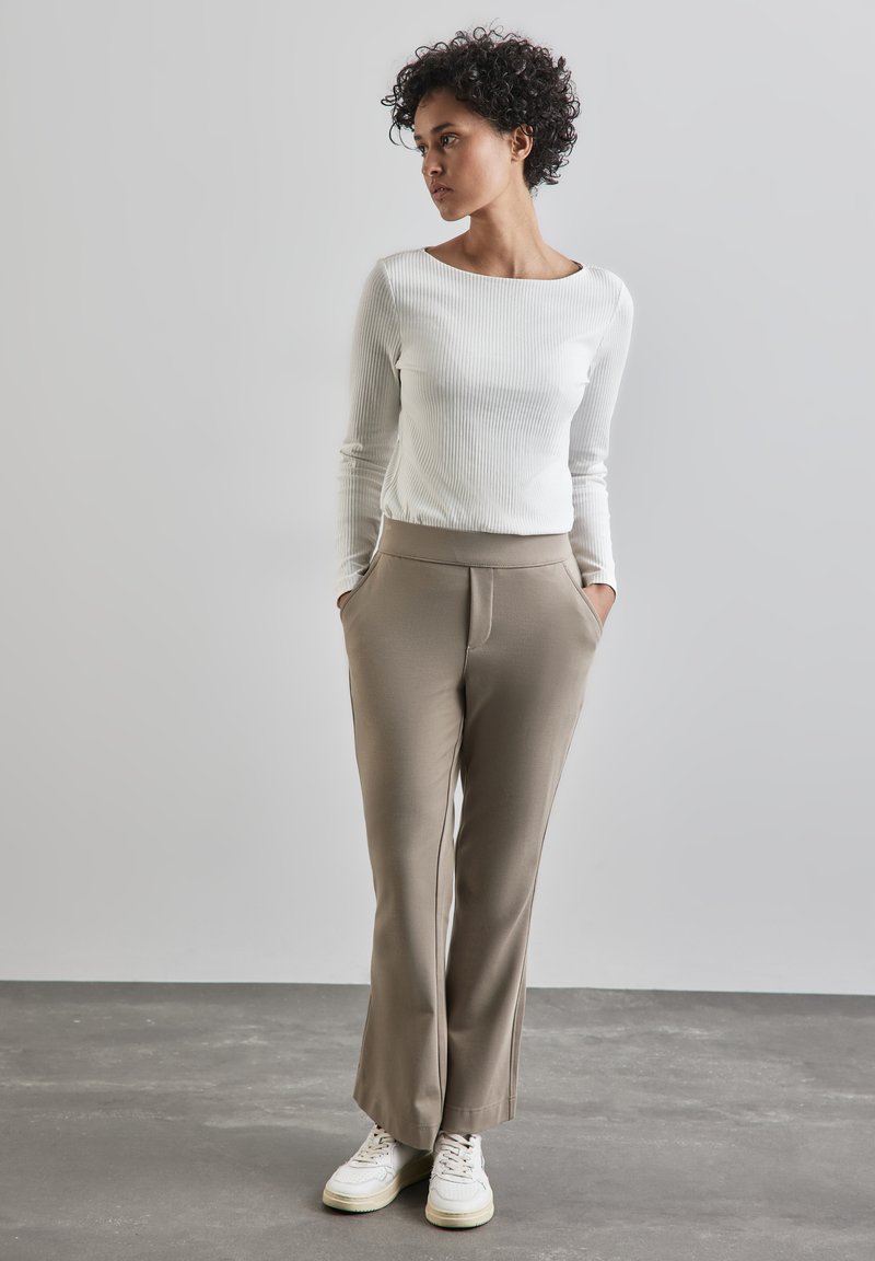 Maglietta bianca a maniche lunghe a costine, pantaloni beige sartoriali con tasche laterali e sneaker bianche. Il modello è in piedi contro uno sfondo grigio chiaro.