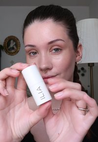 ILIA Make-up-Produkt in einem weißen zylindrischen Tube, der ein einfaches Design mit schwarzer Schrift aufweist. Das Model hält das Produkt in einer gut beleuchteten Umgebung.