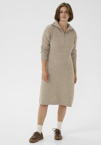 Donna in piedi che indossa un vestito lungo a maniche lunghe a coste beige con colletto a mezza zip, calze bianche e scarpe marroni con lacci su uno sfondo bianco.