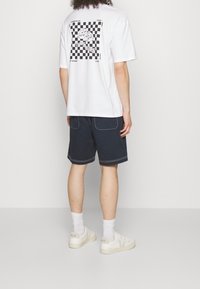 Camiseta blanca con un gráfico en blanco y negro de un patrón de tablero de ajedrez con galletas de dibujos animados. Combinada con pantalones cortos azul oscuro y zapatillas blancas.