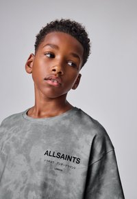 Mladý chlapec s krátkymi kučeravými vlasmi, oblečený v sivom tričku AllSaints, sa pozerá do strany na jednoduchom pozadí.