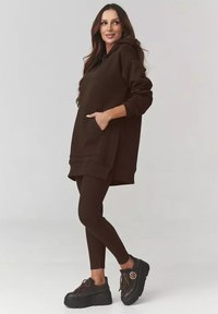 Brązowa, oversize bluza z kapturem i kieszenią typu kangur, zestawiona z brązowymi legginsami o prążkowanej fakturze oraz masywnymi czarnymi butami na platformie. Gładka tekstura, luźny krój.