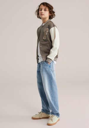 Fiú rendezetlen hajjal, barna és fehér egyetemi stílusú dzsekiben, fehér ingben, bő kék farmert visel, krém színű tornacipőkkel, kezét a zsebében tartva áll.