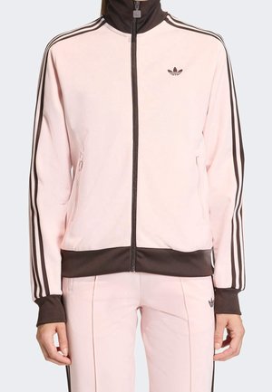 Kobieta w jasnoróżowym dresie Adidas z brązowymi paskami i mankietami, stoi z rękami luźno opuszczonymi wzdłuż ciała, zwrócona przodem.