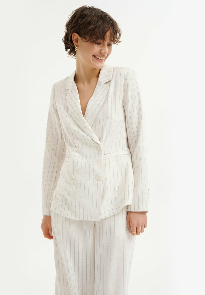 Blazer doppiopetto beige chiaro con righe sottili, caratterizzato da un colletto a rever, due tasche frontali e polsini con bottoni. Tessuto misto lino.