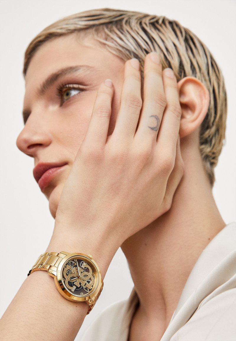 Gold-tone watch with a round face, black patterned dial, and golden hour markers; worn on a wrist with pale skin and a tattoo.