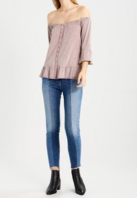 Off-the-shoulder, verouderd roze top met driekwart mouwen en een gerimpelde zoom, gecombineerd met blauwe skinny jeans en zwarte enkellaarsjes.