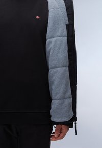 Schwarzer Sweatshirt mit grauem Fleece-Ärmel, ausgestattet mit einem kleinen roten und weißen Flaggenlogo auf der Brust. Strukturierten Stoff und eng anliegende Bündchen.