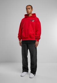 Felpa rossa con cappuccio e cordino, dotata di tasca frontale e con un logo grafico. Indossata con jeans neri larghi e sneakers bianche su uno sfondo neutro.